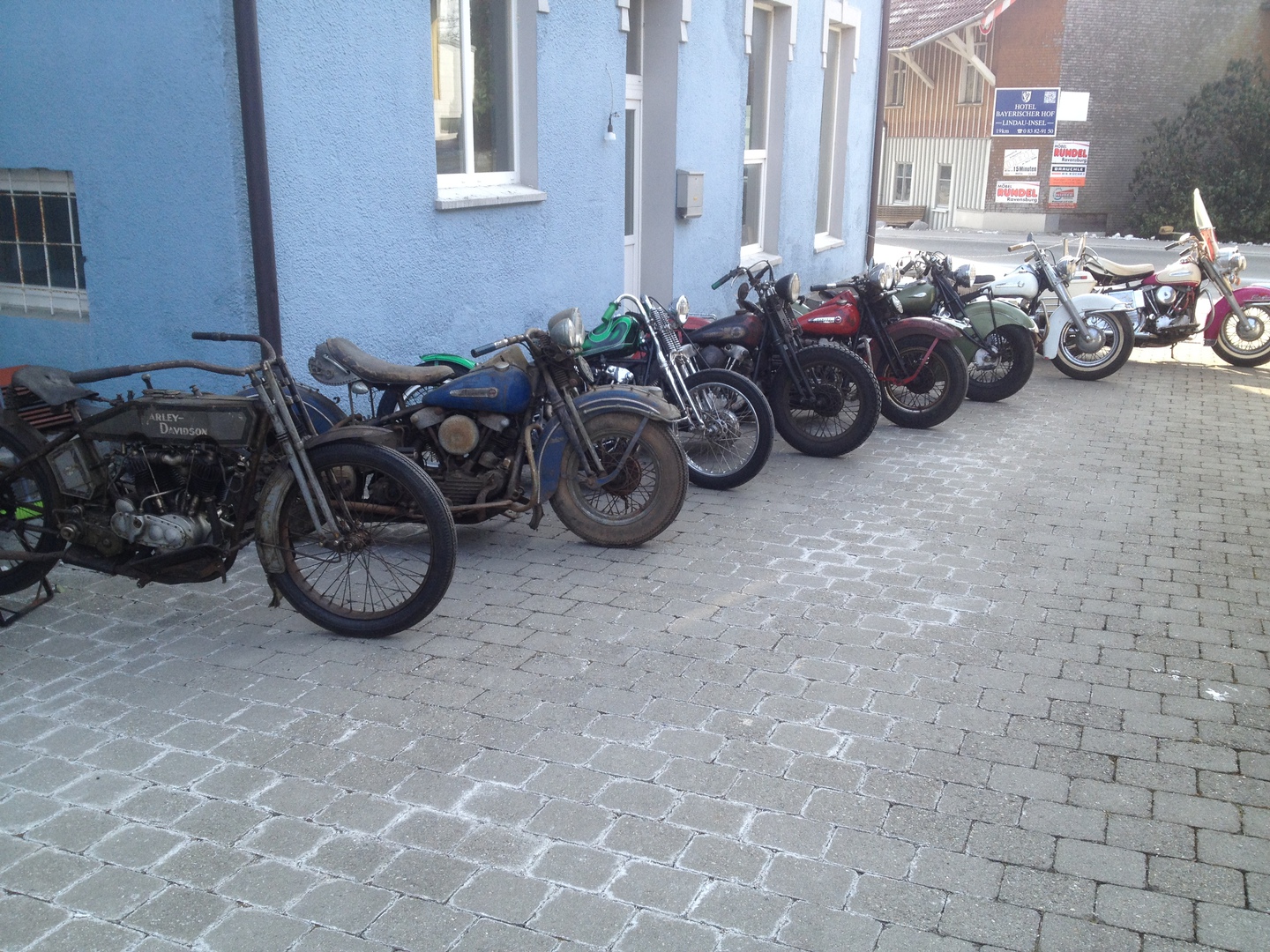 Eine Reihe alter Harleys an der blauen Hauswand, vorn ein Modell aus den Zwanzigern